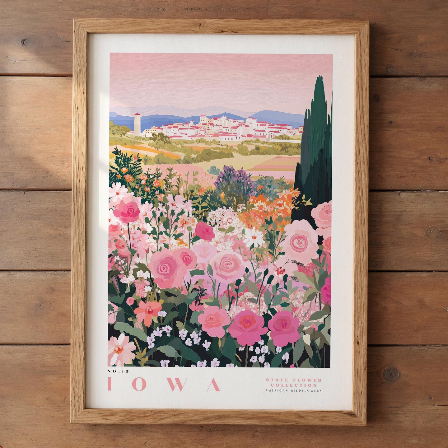 A framed poster depicting a scenic view of Iowa, with a colorful landscape featuring a town, mountains, and a field of flowers.