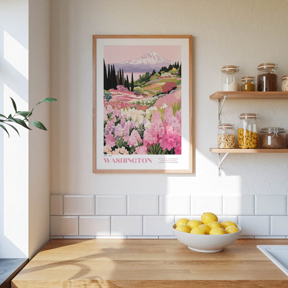 a framed poster of a mountain landscape with pink flowers hanging on a wall above a wooden countertop with a bowl of lemons.