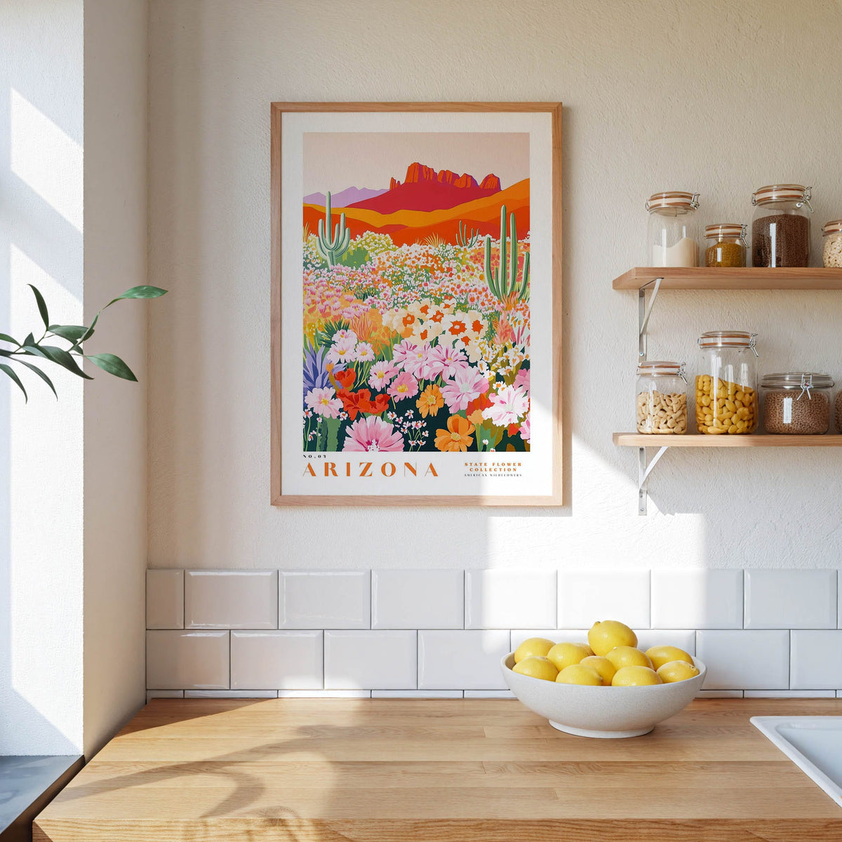 a framed poster of a desert landscape with cacti and flowers, hanging on a wall above a wooden countertop with a bowl of lemons.