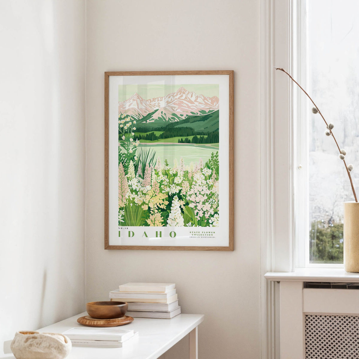 a framed poster of a scenic mountain landscape with a lake and flowers, hanging on a wall in a room with a white table and a window.