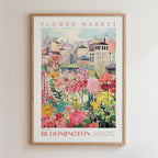 The image is a framed poster depicting a vibrant and colorful flower market in the city of Bloomington, with various flowers and plants in the foreground and a cityscape in the background.