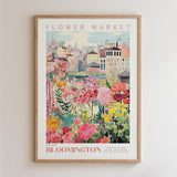 The image is a framed poster depicting a vibrant and colorful flower market in the city of Bloomington, with various flowers and plants in the foreground and a cityscape in the background.