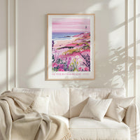 a framed poster of a beach scene with a lighthouse, flowers, and a pink sky, hanging on a wall above a white couch with a blanket draped over it.