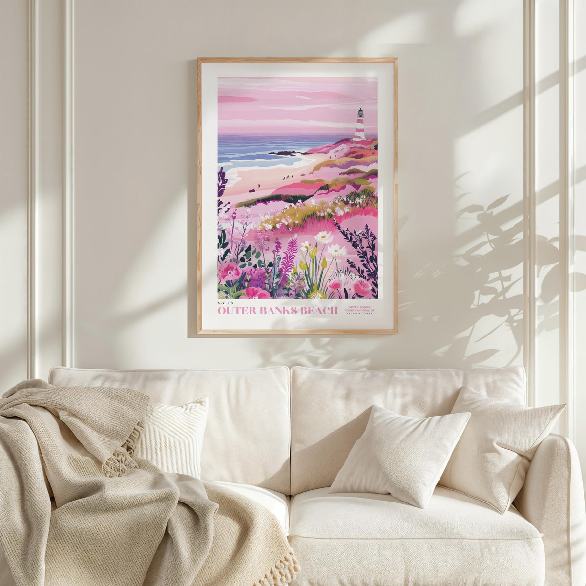 a framed poster of a beach scene with a lighthouse, flowers, and a pink sky, hanging on a wall above a white couch with a blanket draped over it.