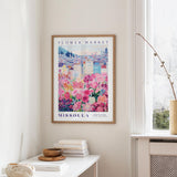 a framed poster of a colorful floral scene hanging on a wall, with a white table and a vase of flowers in the foreground.