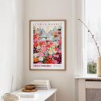 a framed poster of a colorful flower market scene hanging on a wall, with a white table and a vase of flowers in the foreground.