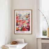 a framed poster of a colorful flower market scene hanging on a wall, with a white table and a vase of flowers in the foreground.