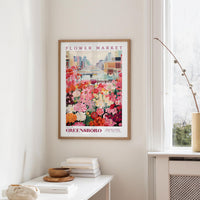 a framed poster of a colorful flower market scene hanging on a wall, with a white table and a vase of flowers in the foreground.