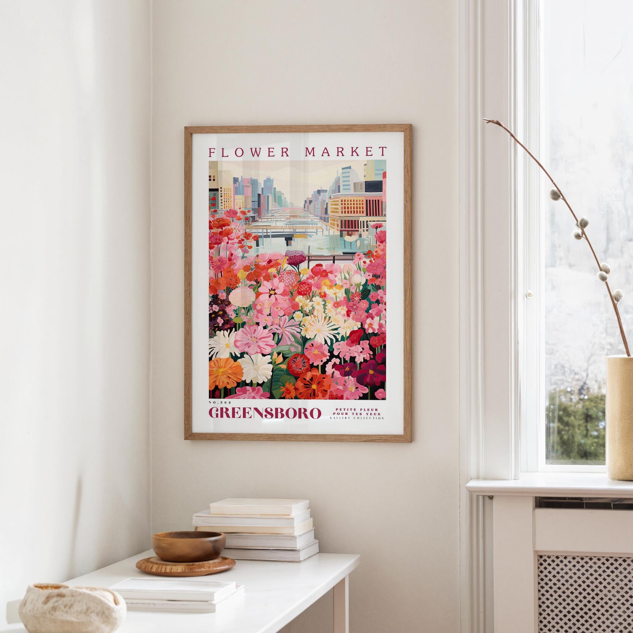 a framed poster of a colorful flower market scene hanging on a wall, with a white table and a vase of flowers in the foreground.