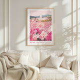 a framed poster of a field of pink flowers hanging on a wall above a white couch with a blanket draped over it.