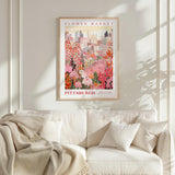 a framed poster of a cityscape with a flower market in the background, hanging on a wall above a white couch with a blanket draped over it.