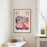 a framed poster of a tropical landscape with palm trees, flowers, and mountains, hanging on a wall in a room with a white table and a window.