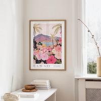 a framed poster of a tropical landscape with palm trees, flowers, and mountains, hanging on a wall in a room with a white table and a window.