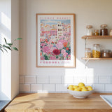 a kitchen scene with a framed poster of a colorful floral market in Cordoba, Spain, hanging on the wall above a wooden counter. On the counter, there is a bowl of lemons and a plant.