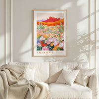 a framed poster of a desert landscape with cacti and flowers, hanging on a wall above a white couch with a blanket draped over it.