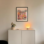 a white cabinet with a potted plant and a lamp on top, set against a wall with a framed poster of a cityscape above it.