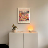a white cabinet with a potted plant and a lamp on top, set against a wall with a framed poster of a cityscape above it.