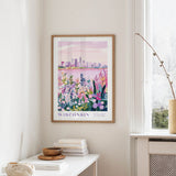 a framed poster of a cityscape with a colorful floral scene, hanging on a wall in a room with a white table and a window.