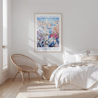 a serene bedroom scene with a bed, a woven chair, and a framed poster of a flower market in Santorini hanging on the wall.