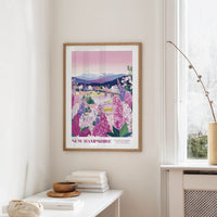 a framed poster of a scenic landscape with mountains, flowers, and a river, hanging on a wall in a room with a window and a small table with books and a vase.