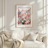 a framed poster of a colorful flower market scene hanging on a wall above a white couch with a blanket draped over it.