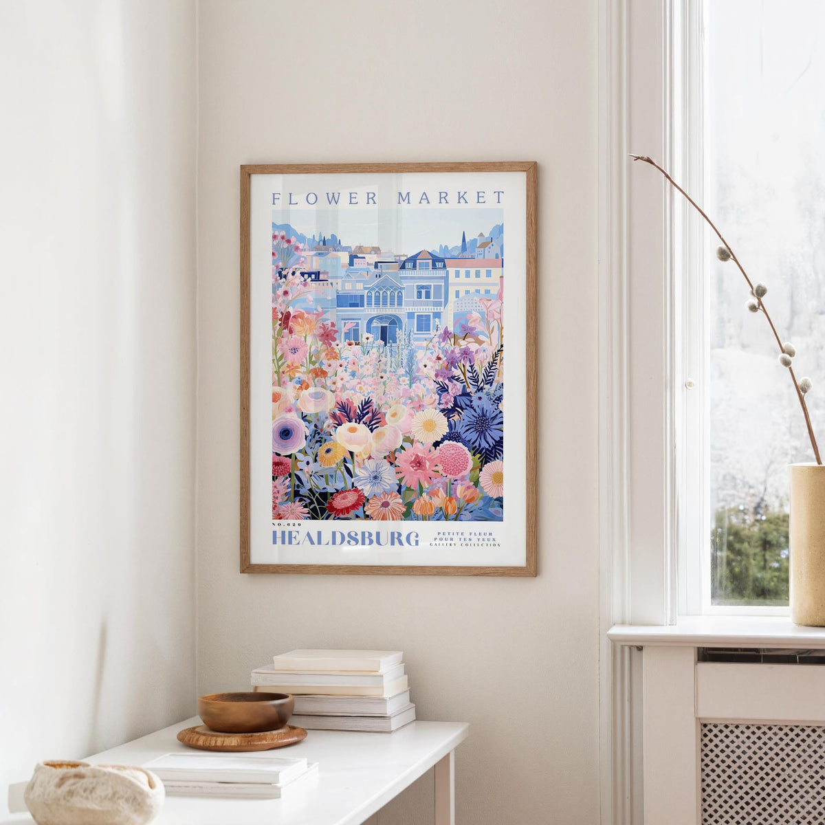 a framed poster of a colorful floral scene hanging on a wall, with a white table and a vase of flowers in the foreground.
