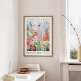 a framed poster of a colorful floral scene hanging on a wall, with a window and a small table with books and a vase in the foreground.