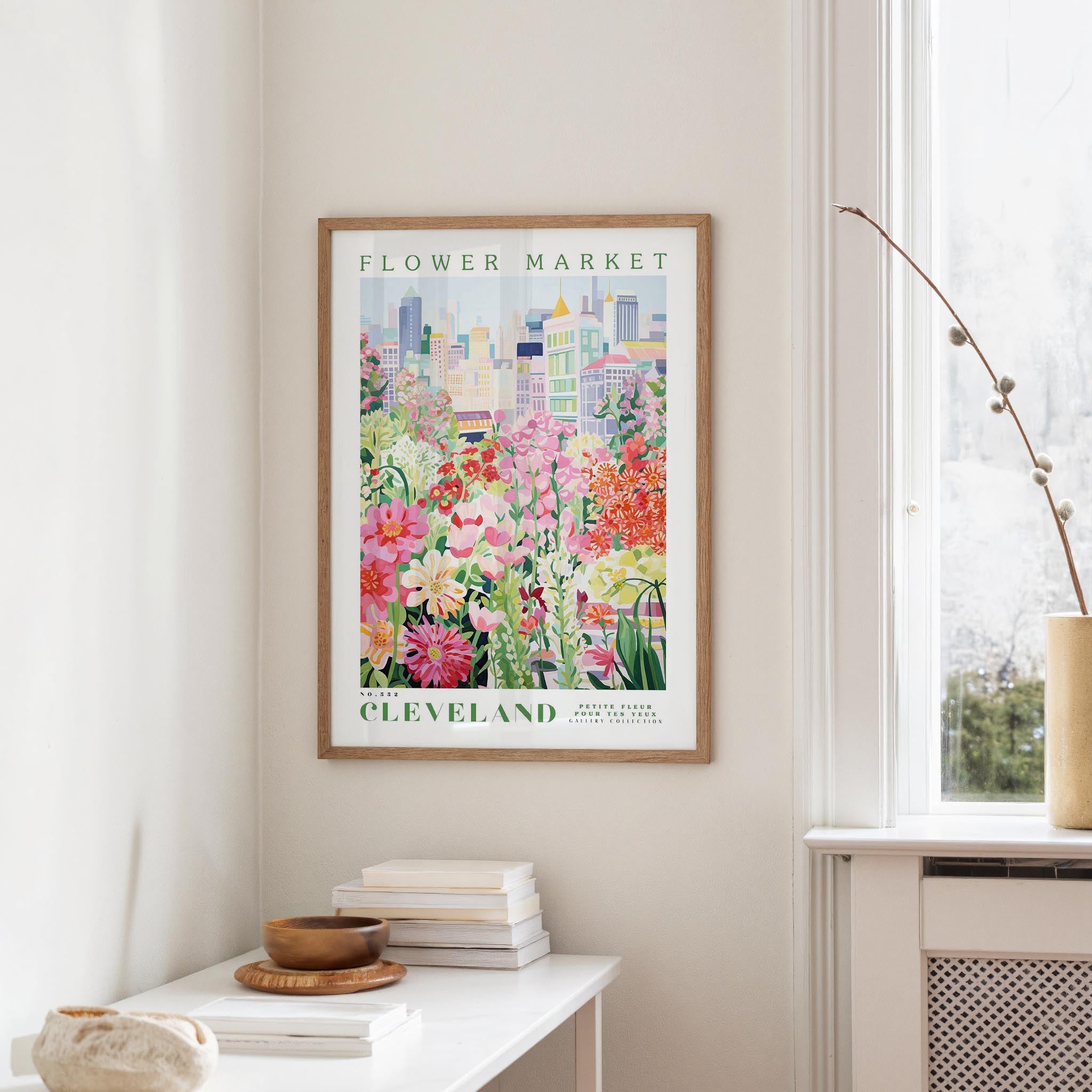a framed poster of a colorful floral scene hanging on a wall, with a window and a small table with books and a vase in the foreground.