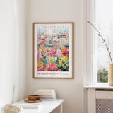 a framed poster of a colorful floral scene hanging on a wall, with a white table and a vase of flowers in the foreground.