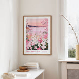 a framed poster of a scenic view of Michigan, with a pink and white floral design, hanging on a wall in a room with a window and a small table with books and a bowl.