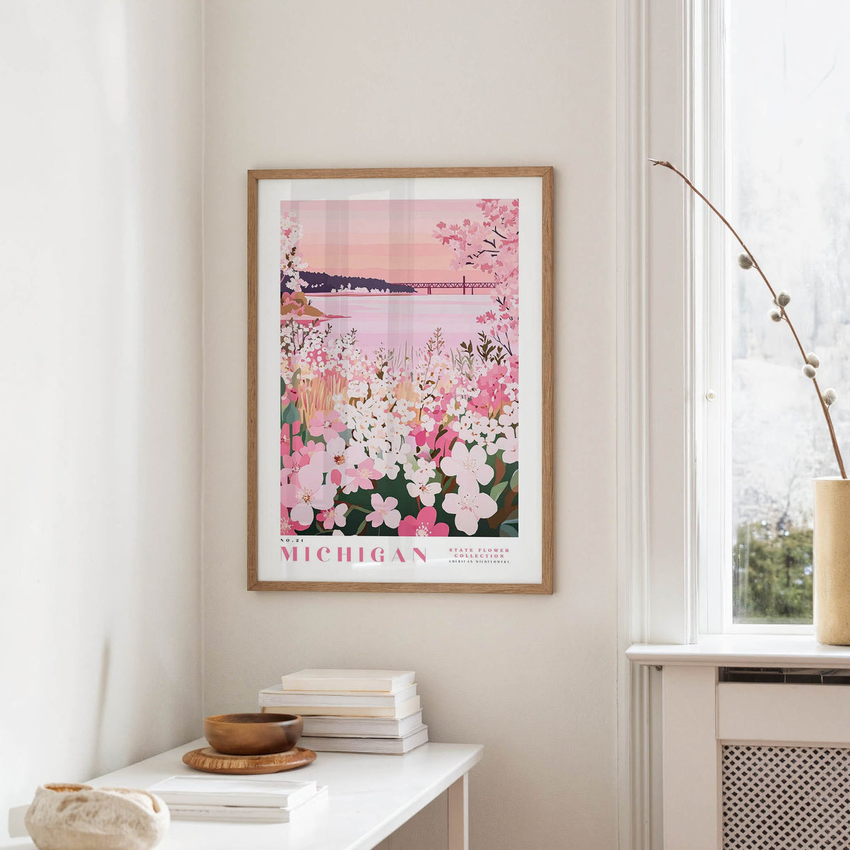 a framed poster of a scenic view of Michigan, with a pink and white floral design, hanging on a wall in a room with a window and a small table with books and a bowl.