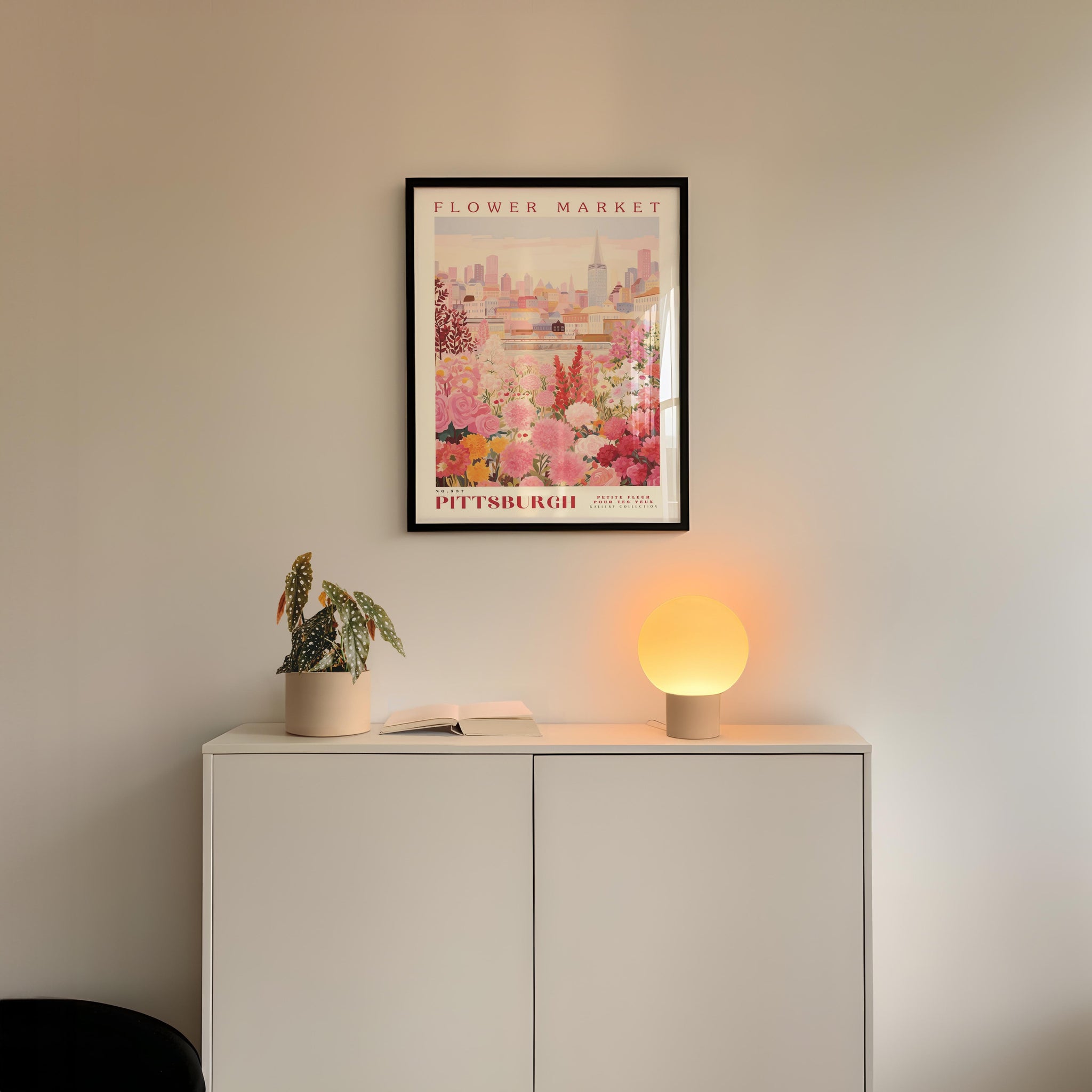 a white cabinet with a potted plant and a lamp on top, set against a wall with a framed poster of a cityscape above it.
