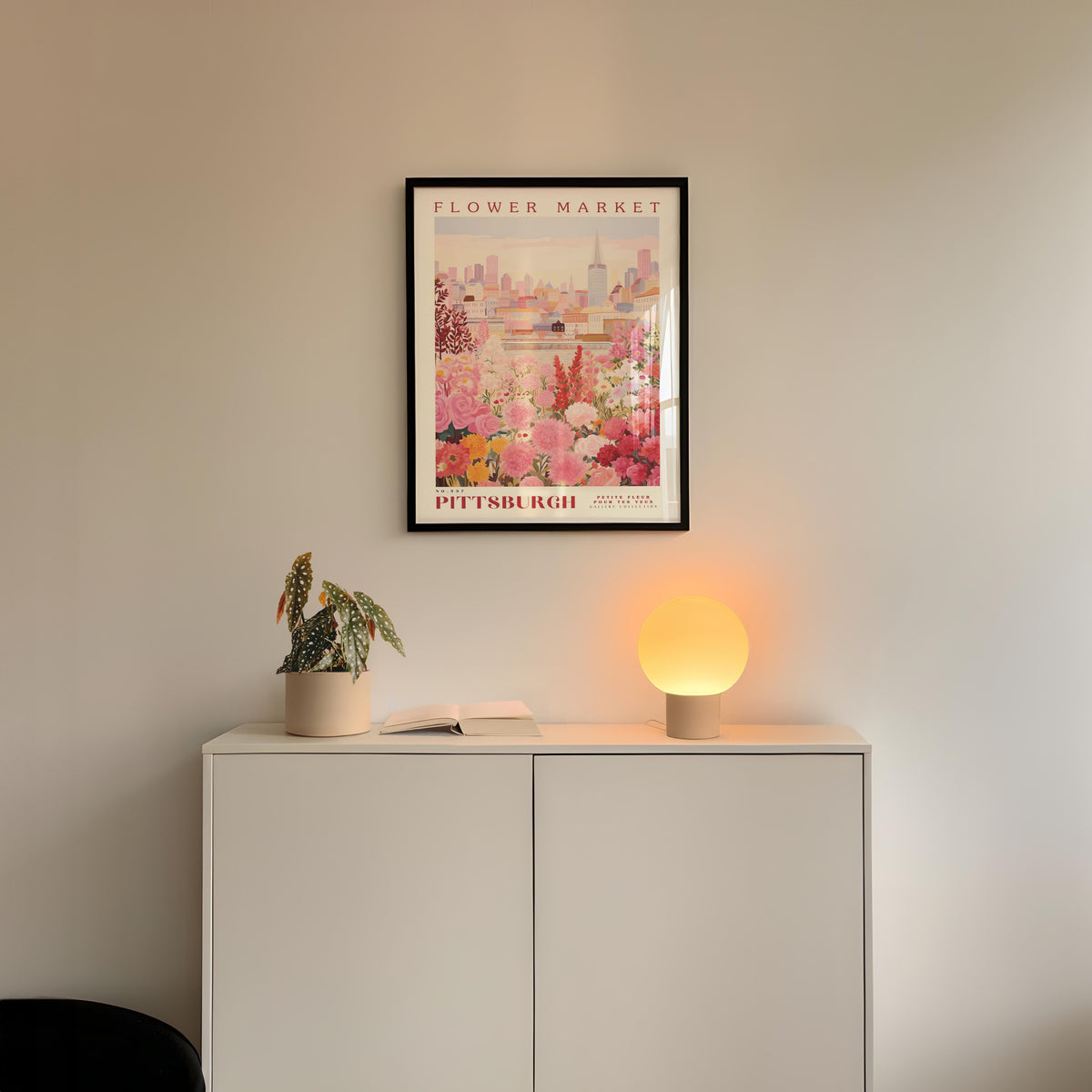 a white cabinet with a potted plant and a lamp on top, set against a wall with a framed poster of a cityscape above it.