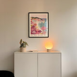 a white cabinet with a potted plant and a lamp on top, set against a wall with a framed poster of a beach scene hanging above it.