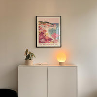 a white cabinet with a potted plant and a lamp on top, set against a wall with a framed poster of a beach scene hanging above it.