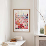a framed poster of a cityscape with a flower market in the background, hanging on a wall in a room with a window and a small table with books and a vase.
