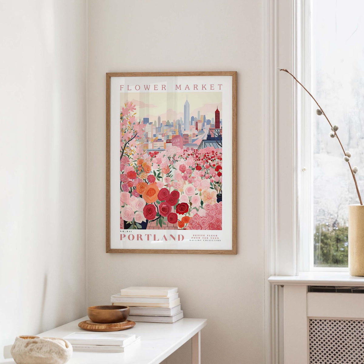 a framed poster of a cityscape with a flower market in the background, hanging on a wall in a room with a window and a small table with books and a vase.