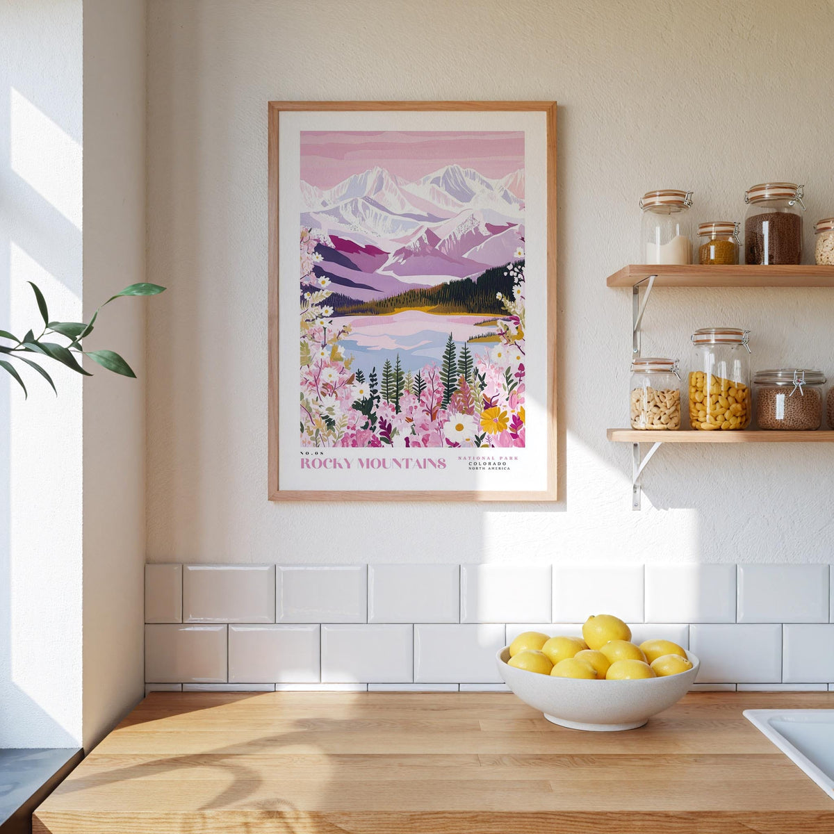 a kitchen counter with a bowl of lemons, a framed poster of Rocky Mountains, and a shelf with jars and bottles.