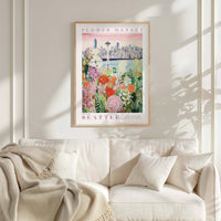 a framed poster of a cityscape with a flower market in the background, hanging on a wall above a white couch with a blanket draped over it.
