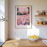 a kitchen scene with a framed poster of a pink and white floral landscape hanging on the wall above a wooden countertop. On the countertop, there is a bowl of lemons and a plant.