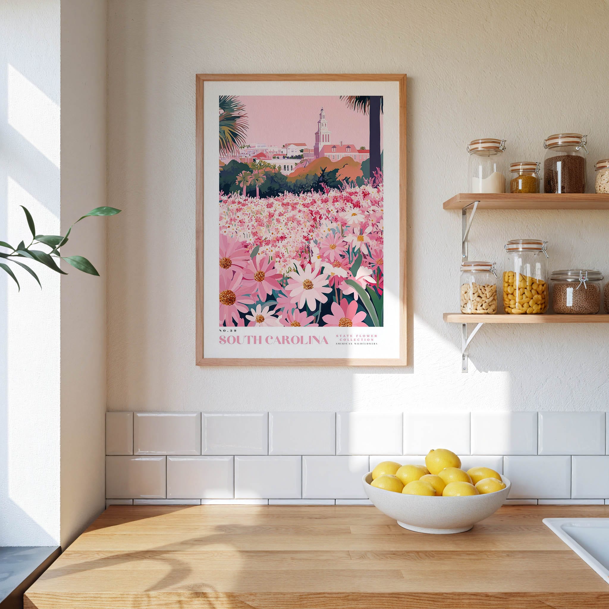 a kitchen scene with a framed poster of a pink and white floral landscape hanging on the wall above a wooden countertop. On the countertop, there is a bowl of lemons and a plant.