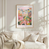 a poster of a colorful cityscape with flowers hanging from the buildings, hanging on a wall above a white couch with pillows.
