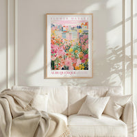 a poster of a colorful cityscape with flowers hanging from the buildings, hanging on a wall above a white couch with pillows.