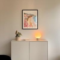 a framed poster of a beach scene hanging on a wall above a white cabinet with a plant and a lamp on top.