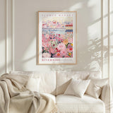 a framed poster of a flower market, with a cityscape in the background, hanging on a wall above a white couch.