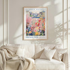 a framed poster of a colorful garden scene hanging on a wall above a white couch with a blanket draped over it.