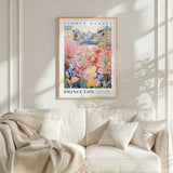 a framed poster of a colorful garden scene hanging on a wall above a white couch with a blanket draped over it.
