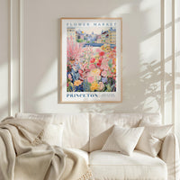 a framed poster of a colorful garden scene hanging on a wall above a white couch with a blanket draped over it.