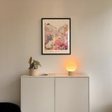 a white cabinet with a potted plant and a lamp on top, set against a wall with a framed poster of a colorful cityscape above it.
