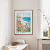a framed poster of a beach scene with a colorful, tropical landscape, hanging on a wall in a room with a white table and a vase of flowers.